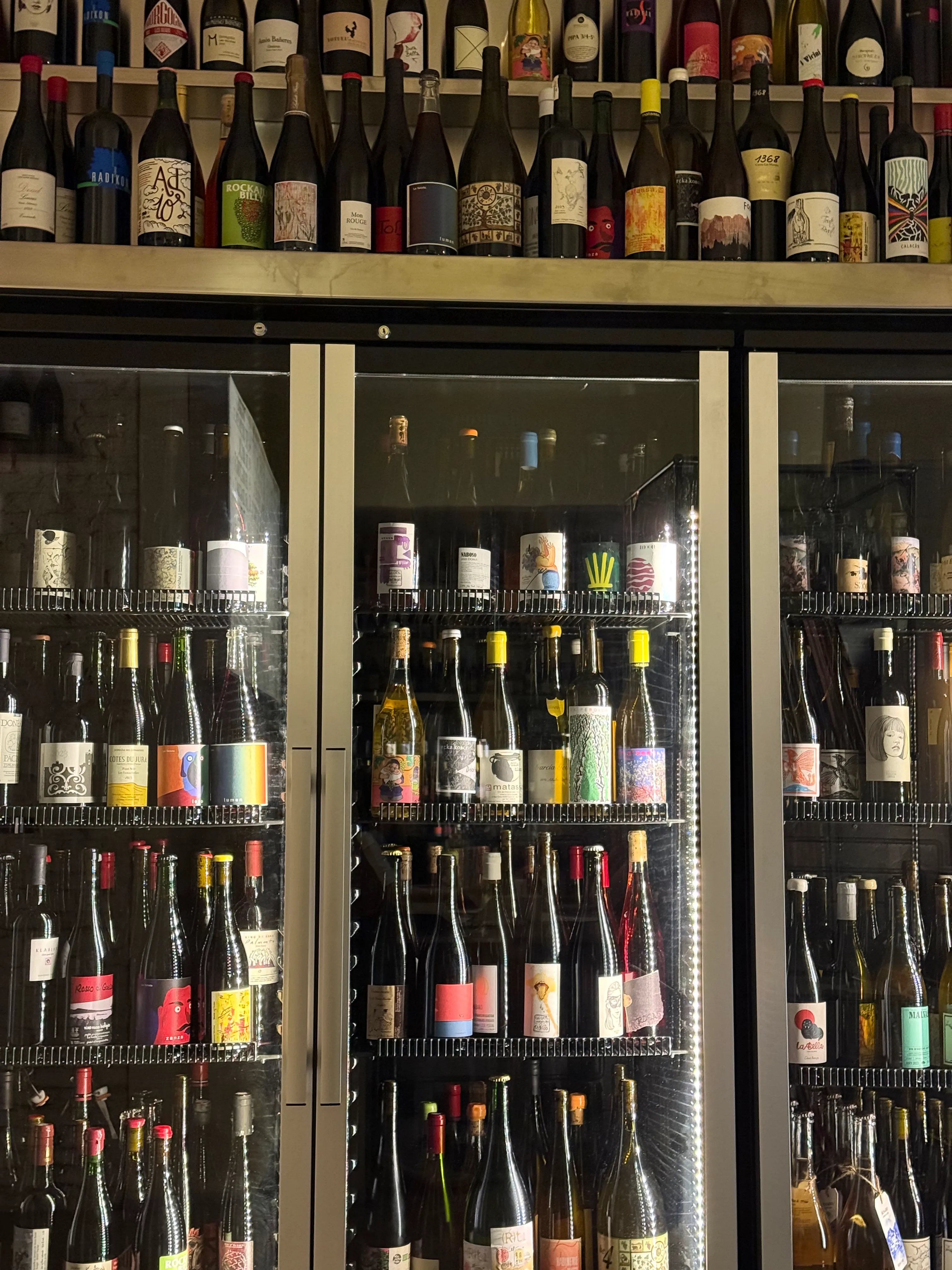 Pared de botellas de vino y una nevera de tres puertas llena en un wine bar, con las estanterías abarrotadas de referencias de suelo a techo.