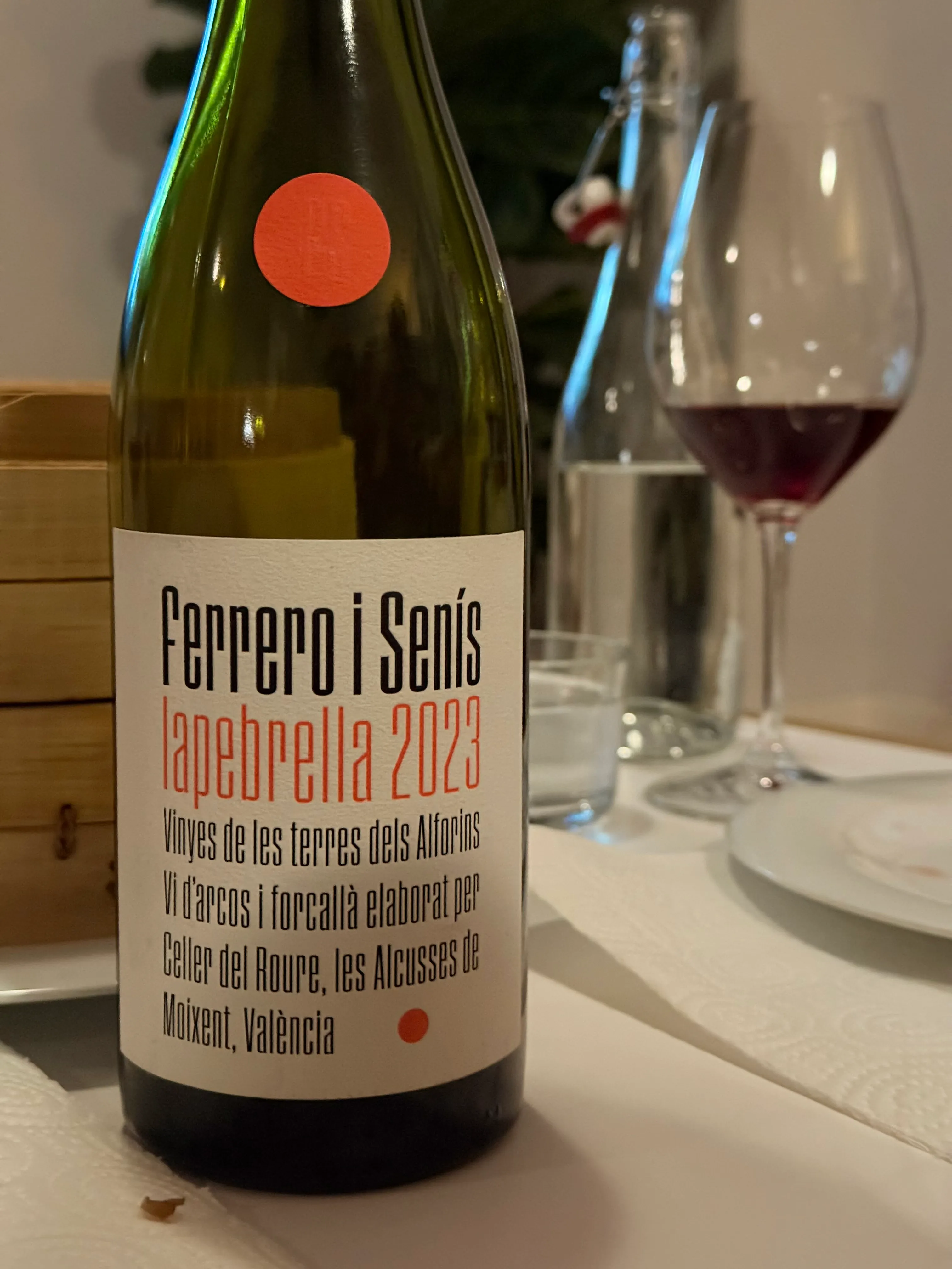 Botella de La Pebrella 2023 de Celler del Roure, de la colección Ferrero i Senís, sobre un mantel blanco junto a una copa de tinto servida.