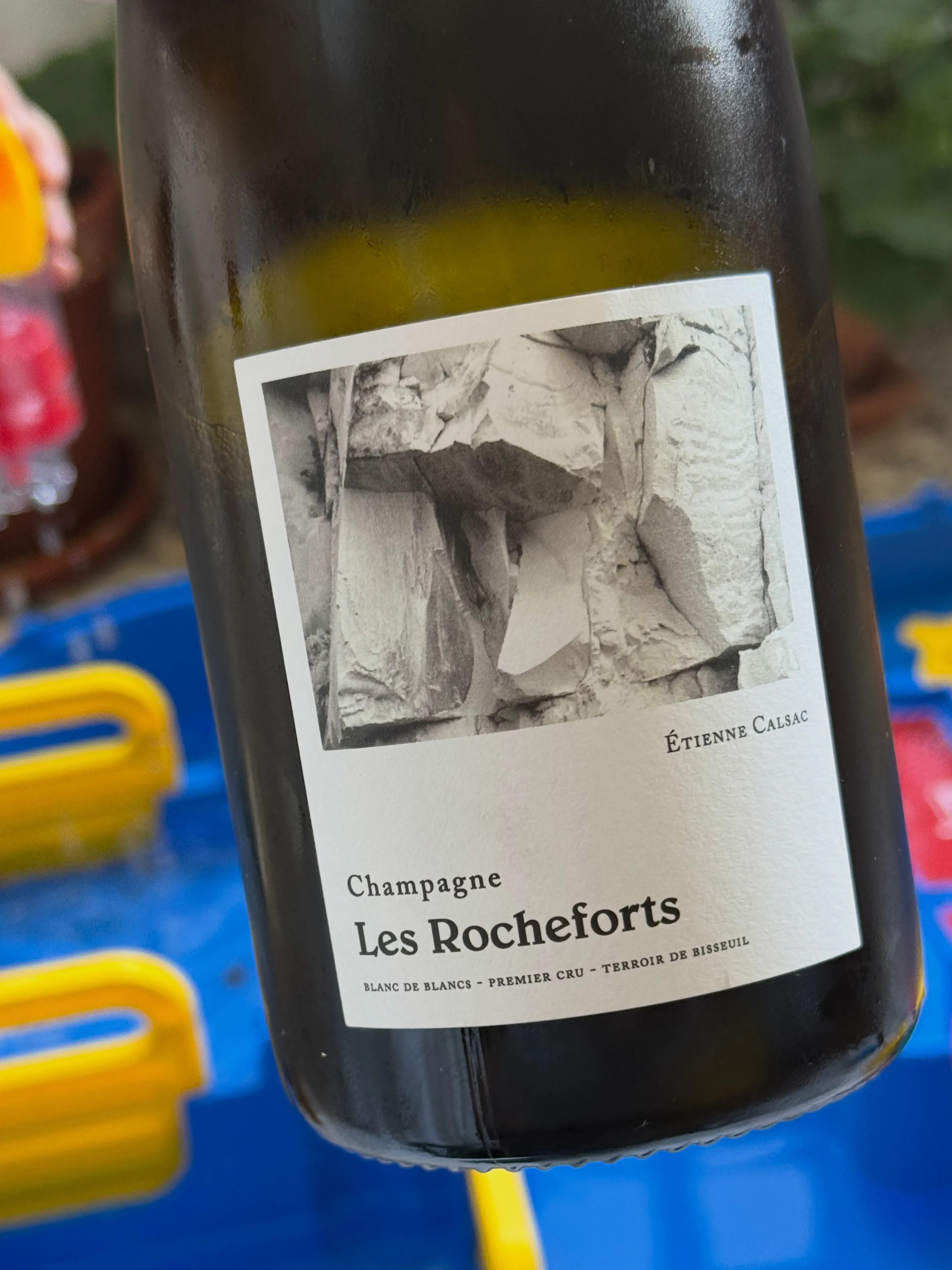 Botella del champán Les Rocheforts de Étienne Calsac, Blanc de Blancs Premier Cru de Bisseuil, con su etiqueta en blanco y negro de una roca de tiza, sobre una mesa de exterior de colores.