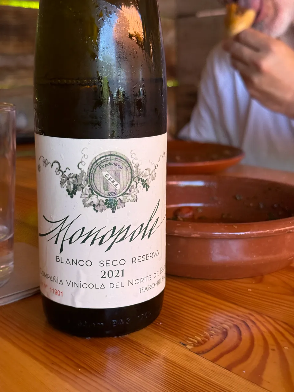 Botella del Monopole Blanco Seco Reserva 2021 de CVNE, con su etiqueta clásica y condensación de venir bien fría, sobre una mesa de madera junto a una cazuela de barro.
