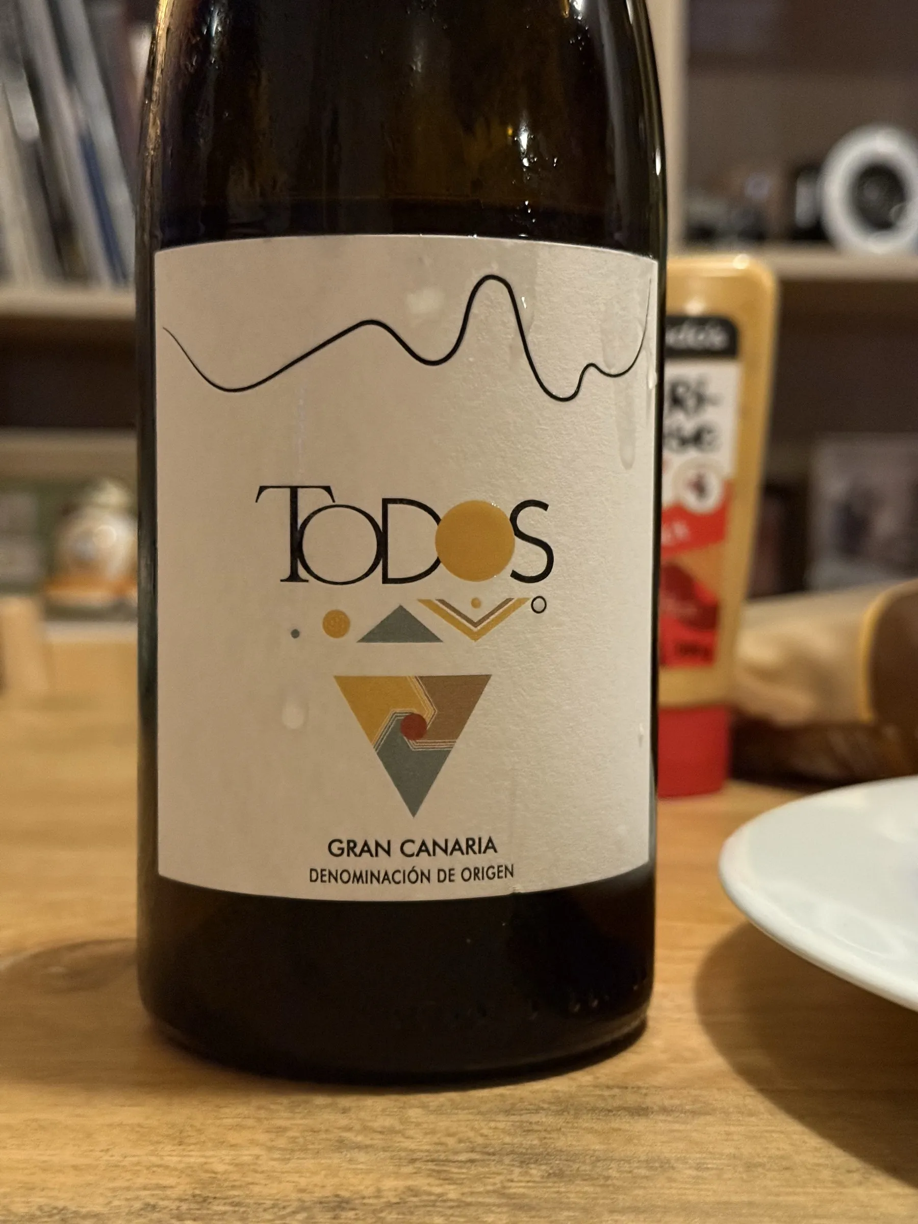 Botella del Todos Blanco 2024 de Gran Canaria sobre una mesa de madera, con la etiqueta blanca del triángulo invertido y el sello de la Denominación de Origen Gran Canaria.