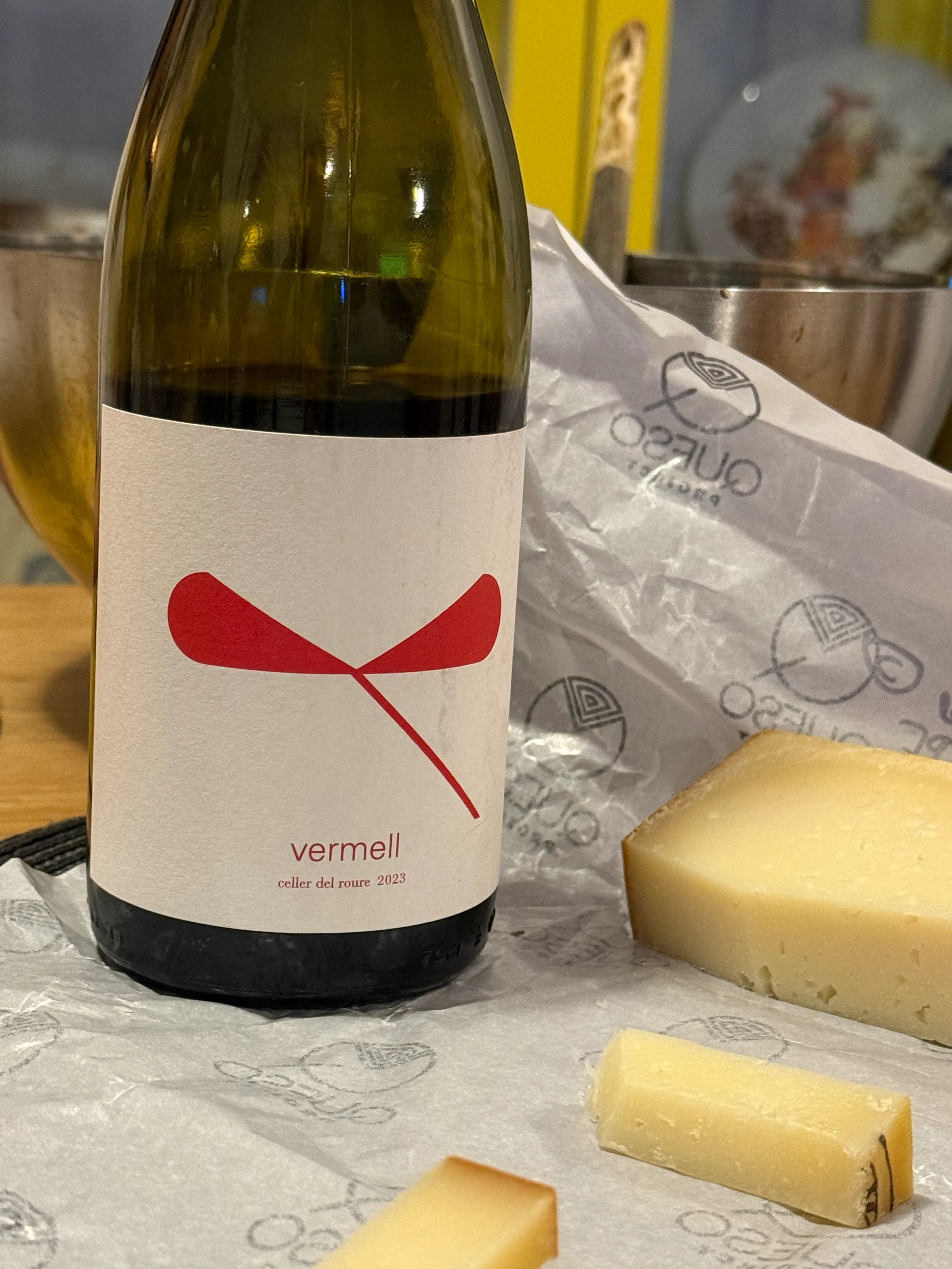Botella de tinto Vermell de Celler del Roure tumbada junto a varias cuñas de queso de El Queso Project sobre el papel de la quesería.